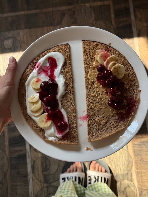 A hand holds a white plate with two crepes topped with yogurt, banana slices, and a berry compote, viewed from above alongside feet wearing Nike slides on a tiled floor.