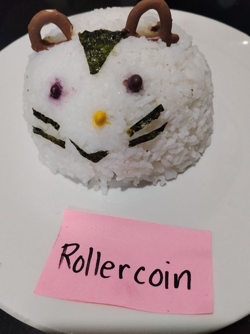 A rice ball art piece shaped like the same cartoon bear face with nori and chocolate details, served on a white plate with a pink 'Rollercoin' sticky note.