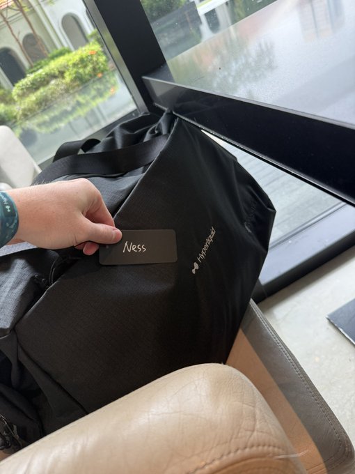 A hand holds a name tag reading "Ness" against a black Hyperliquid-branded bag, suggesting the owner is attending a Hyperliquid event. The casual setting, with the bag resting on a couch near a window, hints at a relaxed networking or conference gathering hosted by the crypto platform.