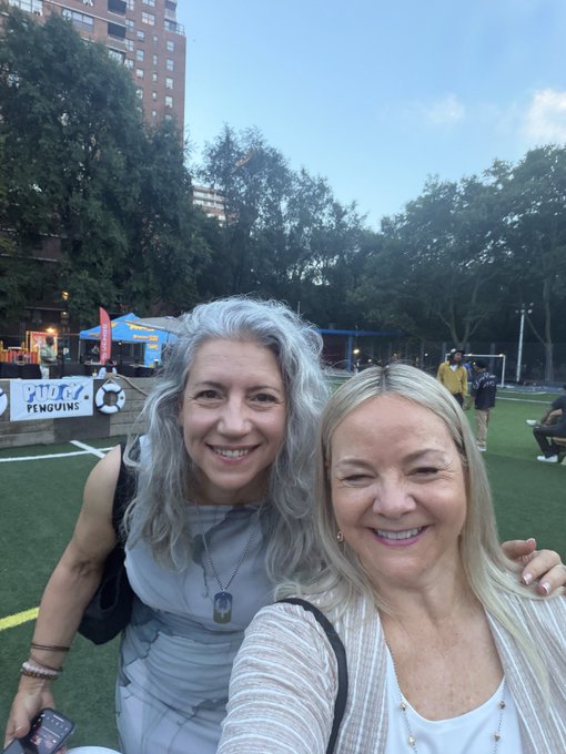 Two smiling women taking a selfie outdoors at what appears to be a sports field, with 'PENGUINS' signage and food trucks visible in the background.