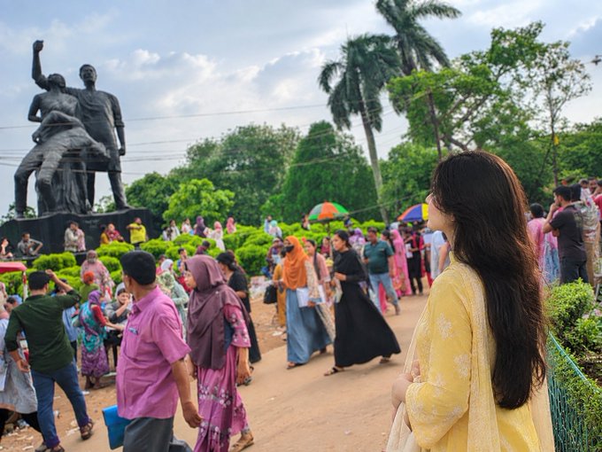 A woman with long dark hair in a yellow dress stands looking at a bronze statue in a park surrounded by palm trees and crowds of people.