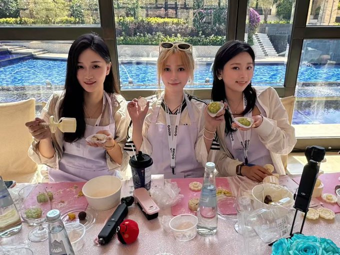 Three young women wearing white shirts and pink aprons sit at a table making desserts, holding small pastries and a piping bag, with a pool visible through the windows behind them.