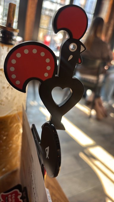 A decorative metal rooster-shaped beer tap handle, likely featuring a brand logo, is mounted on a wooden bar in what appears to be a cozy restaurant or café setting with patrons seated in the background. The playful rooster design and rustic ambiance suggest the location is a craft beer establishment or gastropub, fitting the playful "Guess where I am" caption.