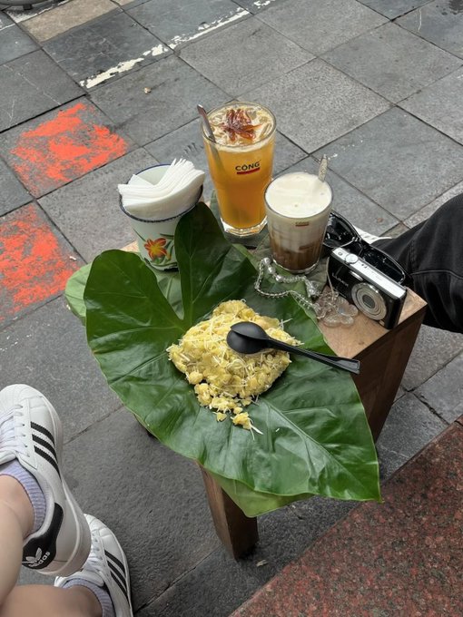 A meal of rice served on a large green leaf sits on a small wooden stool alongside two iced drinks—one in a glass branded "CONG"—and a vintage camera, with a pair of white Adidas sneakers visible in the foreground. The scene captures a casual breakfast moment that contrasts with the thoughtful caption about prioritizing real infrastructure over fleeting narratives in crypto.