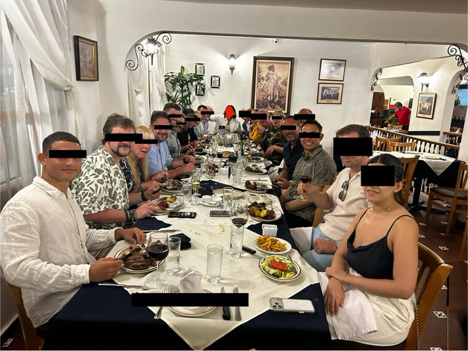 A group of people sit together around a long table at a steakhouse in Panama, enjoying a meal with wine glasses, plates of steak and fries, and smartphones scattered across the tablecloth, with faces redacted for privacy. The image ties to the post's playful theme of keeping identities hidden due to insider knowledge about XRP liquidity.