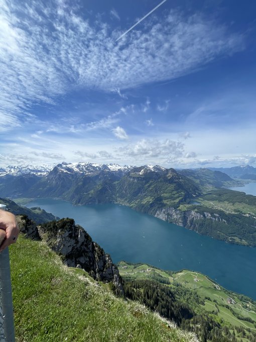 A scenic mountain vista photo taken from a high vantage point overlooking a turquoise alpine lake surrounded by green valleys and snow-capped peaks under a blue sky with wispy clouds.