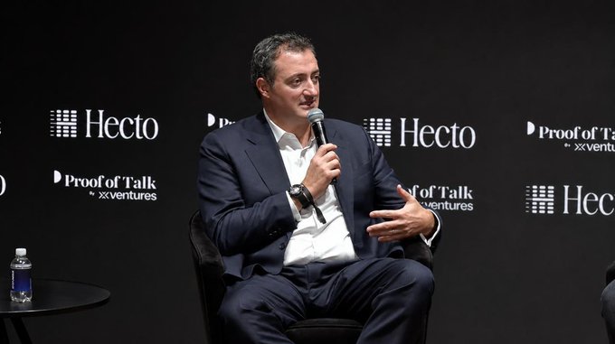 The image features a man sitting in a chair, holding a microphone and speaking into it. He appears to be at an event or conference, possibly addressing an audience. The background of the image shows a black wall with white text that reads "ProfitOfAdventure" and "HECTO". There is also a water bottle placed on the table next to the man.