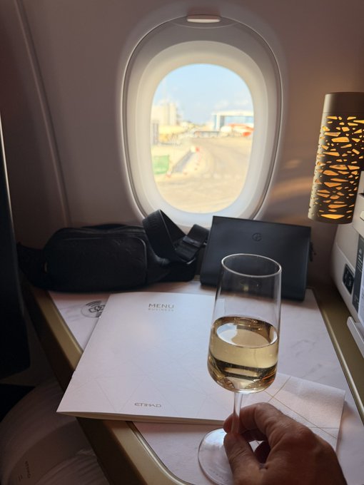 A hand raises a glass of champagne in a toast aboard an Etihad business class flight, with a menu and amenity kit visible on the tray table. The image illustrates the post's celebration of upgrading to business class, likely made possible by Bitcoin profits.