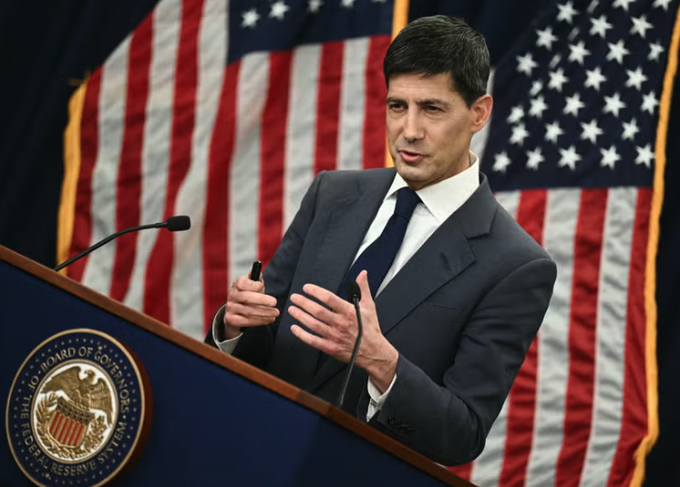 A man in a dark suit and tie stands at a podium bearing the seal of the Federal Reserve Board of Governors, speaking with his hands raised in a gesture, with American flags visible in the background—suggesting this image was taken during a Federal Reserve press conference related to the FOMC's hawkish rate hold decision.