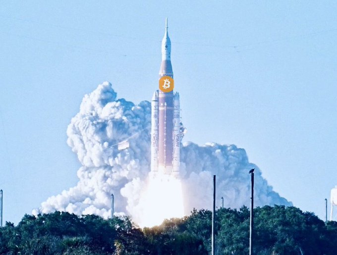 A Saturn V rocket lifts off amid billowing smoke, with a Bitcoin logo digitally edited onto its side—visually punning on the phrase "To the moon" to depict the cryptocurrency's meteoric rise.