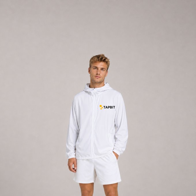A young man with blond hair models a white windbreaker jacket and white shorts against a plain gray background; the jacket bears the logo 'TAPBIT'.