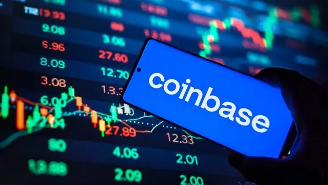 A smartphone displaying the "coinbase" logo is held up against a background of a stock market screen showing colorful trading charts and various price numbers like 27.89, 12.33, and 27.60, visually reinforcing the article about Coinbase's expansion into AI-powered financial infrastructure and trading tools.
