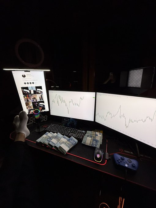 A nighttime photo of a trading desk setup with three monitors displaying stock/crypto charts, a keyboard, bundles of cash (euros), a gaming controller, and a small cat figurine on the desk.
