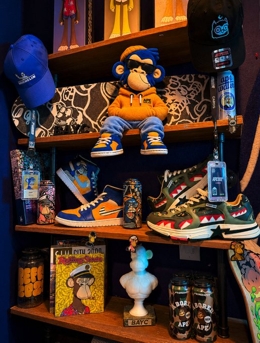The image features a shelf with various items, including a stuffed monkey toy, skateboard, and several cans of beer. There are also two books displayed on the shelf. The main focus of the image is the collection of items, which includes a stuffed monkey, a skateboard, and some cans of beer. The presence of these items suggests that they might be part of an event or gathering related to the post "GM BAYC".