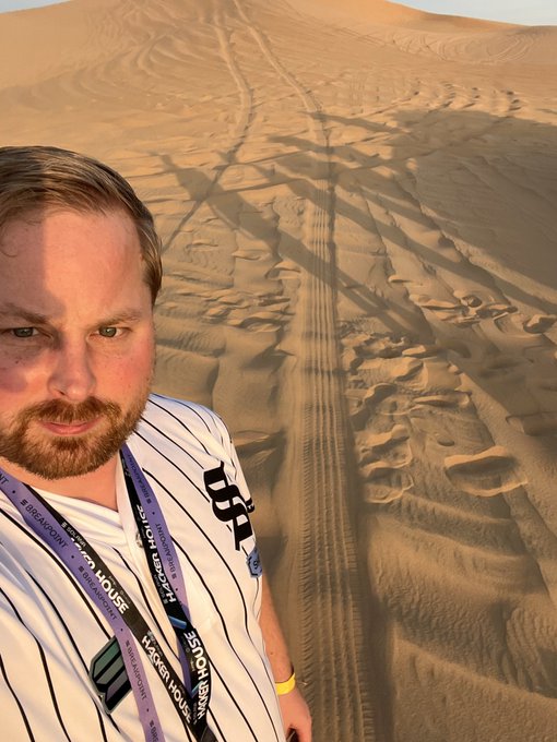 The image features a man standing in a desert with his hands on his hips, wearing a baseball jersey. The sandy terrain stretches out behind him, leading towards an unseen horizon line. The photo captures the vastness of the desert landscape, emphasizing its expansive nature. In the background, there are tracks left by vehicles or people that have passed through the area, indicating human presence in this remote location.