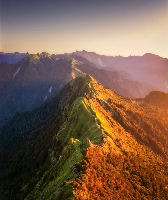 An aerial view of a majestic mountain ridge with green slopes on one side and autumn orange foliage on the other, bathed in warm golden sunset light.