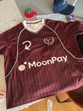 A maroon soccer-style jersey with "MoonPay" prominently displayed across the chest and the number "2" on the upper left, shown alongside a small strawberry plush and other accessories, showcasing the high-quality merchandise that the poster praises in their caption.