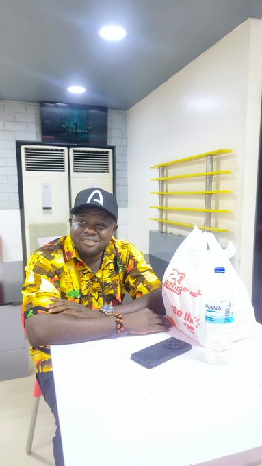 The image features a man sitting at a table wearing a yellow shirt and a baseball cap. He has his arms crossed over his chest while posing for the picture. On the table, there are two water bottles - one near him and another further away from him. The background of the photo shows a wall with shelves on it.