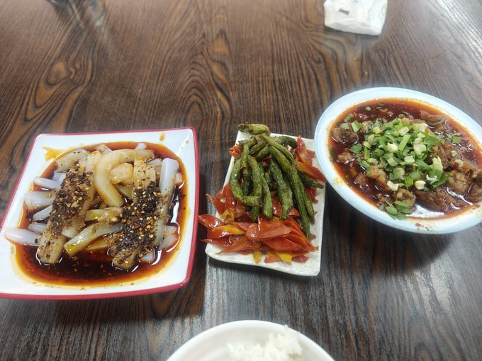 Three Chinese dishes are displayed on a wooden table: sliced noodles with chili oil and sesame seeds, stir-fried green beans with peppers, and a meat dish garnished with scallions in red sauce, with a bowl of rice partially visible.
