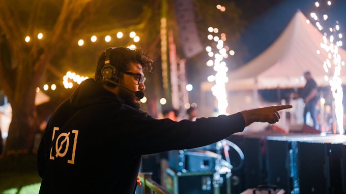 A man wearing headphones and a black sweatshirt with "[Ø]" printed on it points across a stage at night, with string lights and a white tent in the background of an outdoor event. The image captures a behind-the-scenes moment of the music festival mentioned in the caption, showing the organizer directing the festivities.
