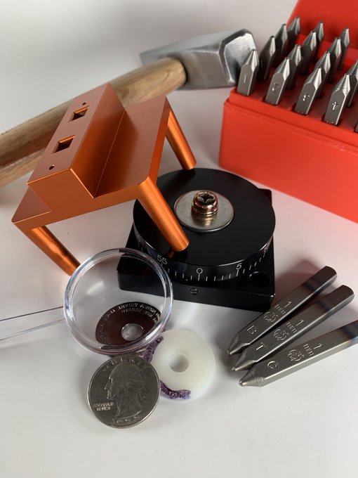 A still life of coin stamping tools including a hammer, a red case of letter/number punches, metal coin dies, a magnifying loupe, and a US quarter used to test size.