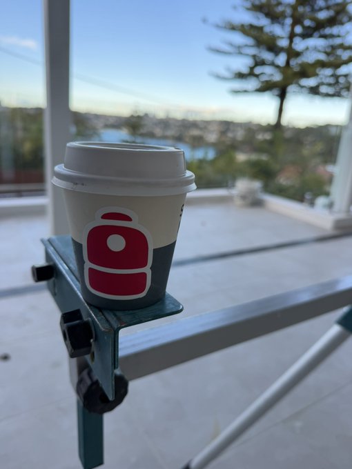 A small takeaway coffee cup with a white lid sitting on a glass railing, with a view of a coastal city and trees in the background.