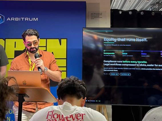 A man with glasses and a beard speaking into a microphone at an Arbitrum event, with a screen behind him displaying 'Equity that runs itself' and information about the token lifecycle.