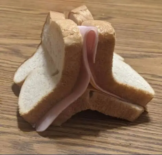 A sandwich made with sliced white bread and ham has been arranged in a complex, gravity-defying structure on a wooden surface, showcasing an unusually creative and intricate assembly.