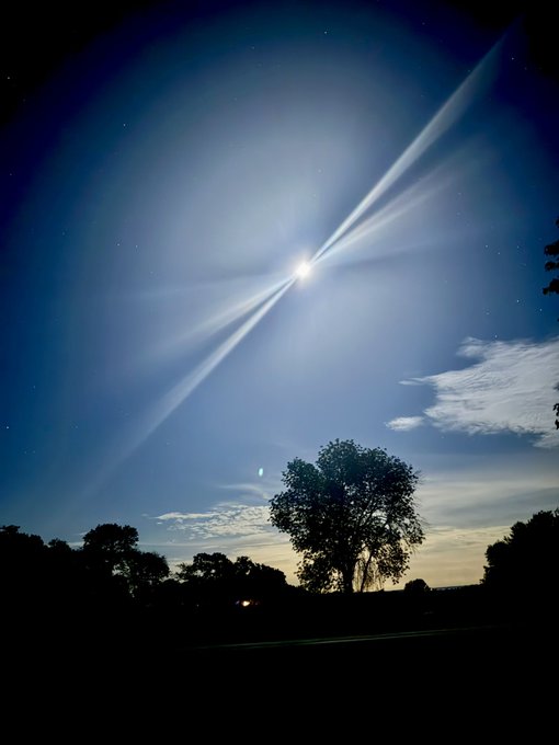 A nighttime photograph of a brilliantly bright moon creating dramatic light rays in a starry blue sky, with the silhouettes of trees along the horizon.