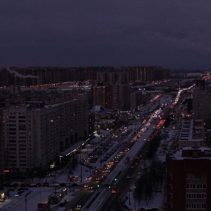 An aerial nighttime view of a snow-covered city with a long avenue of traffic, streetlights, and illuminated high-rise apartment buildings under a dark sky.