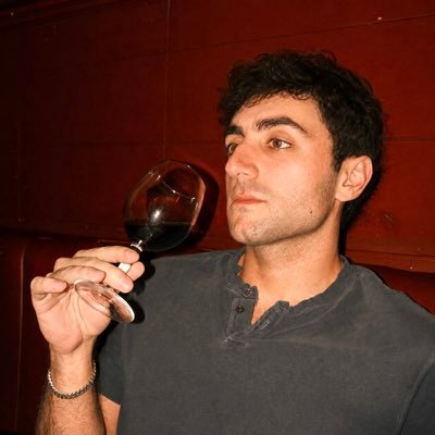 A man with dark curly hair wearing a dark grey henley shirt, holding a glass of red wine near his face while looking to the side against a red paneled background.