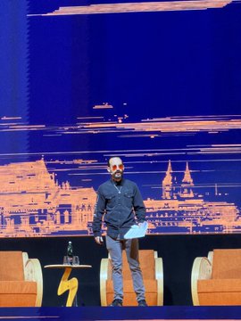 The image features a man standing in front of a blue wall with orange chairs arranged behind him, giving off an impression that he's speaking at an event or conference. The background includes a city skyline, suggesting that this could be taking place in a large urban setting like Prague. The man is wearing sunglasses and holding a piece of paper, possibly a presentation or speech. The image also contains text elements such as the words "@BTCPrague" and "@francispouliot", which may provide additional context about the event or location where this scene takes place.