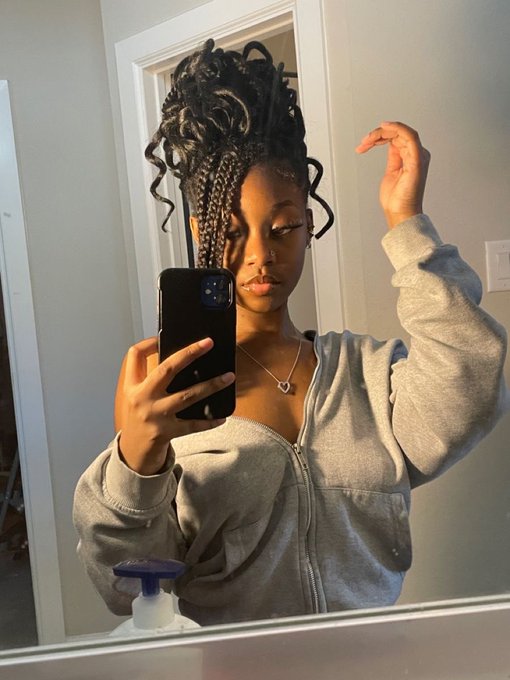The image features a woman taking a selfie with her cell phone in front of a mirror. She appears to be wearing a grey sweatshirt, which complements the blue bottle she has placed next to the mirror. The background seems to be a bathroom setting, as there is a sink visible in the scene. The post includes text that reads "Good morning happy Friday no work for me today, I might sleep off after this observe $RIVER and enjoy your beautiful day".