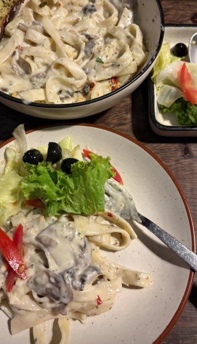 A photo of a meal consisting of a creamy mushroom pasta dish served in a white bowl with a side salad of lettuce, tomato, and black olives on a plate, with a fork resting on the food.