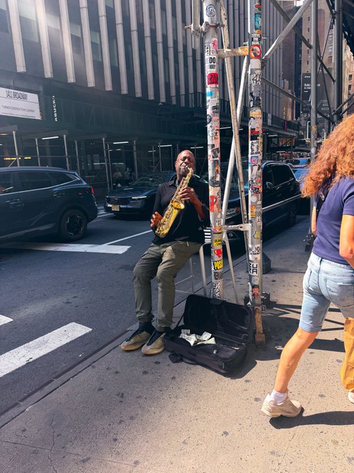 A street musician plays a saxophone on a city sidewalk, leaning against a pole covered in stickers, with his open instrument case on the ground for tips.