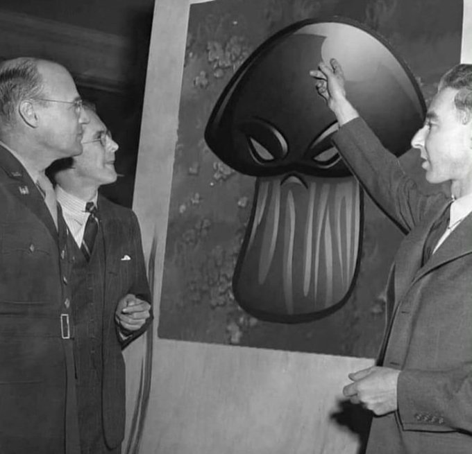A black-and-white historical photo shows two men in military uniform observing as another man points to a large painting of a mysterious figure with glowing eyes and a tentacled face.