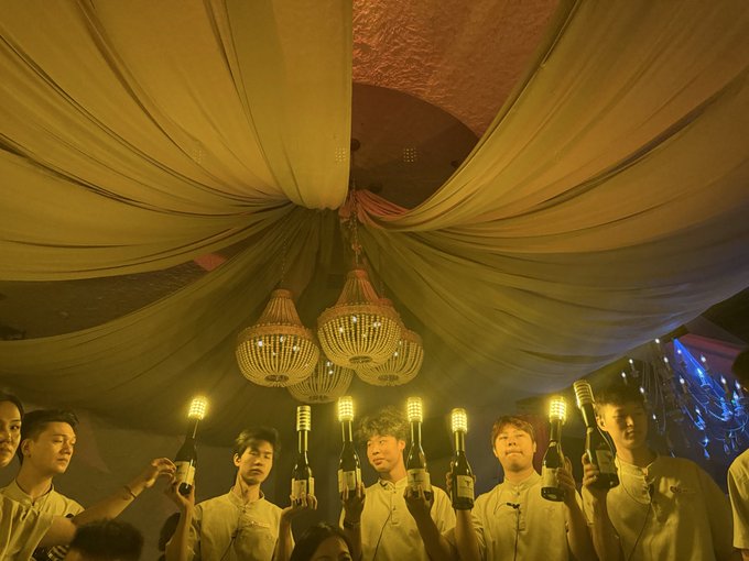 A group of men in white shirts holding up lit bottle lamps under draped fabric and chandeliers.