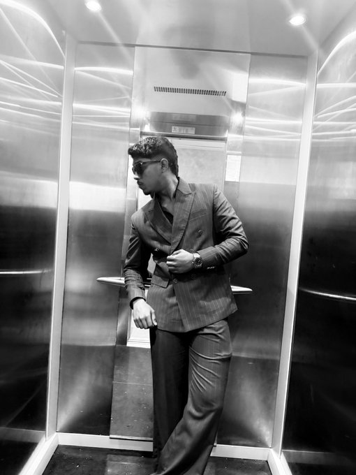 A black and white photograph of a man in a pinstripe suit and sunglasses posing inside a metallic elevator.