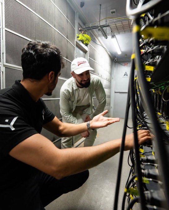 Two men in a server room examining equipment; one wears a black shirt while the other wears a striped shirt and a 'Budweiser' cap.
