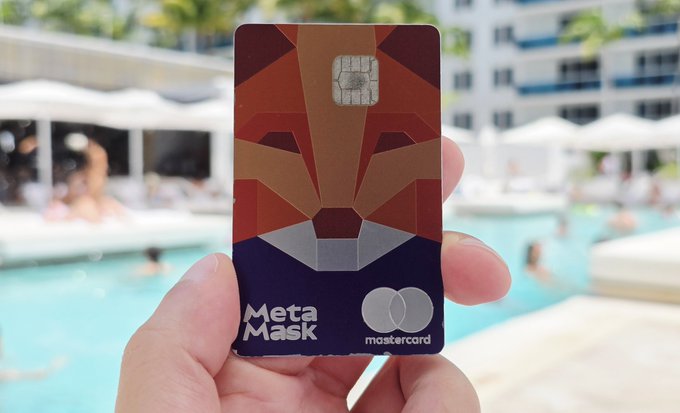 A hand holding a MetaMask Mastercard debit card with a geometric fox design, shown against the backdrop of a swimming pool area.