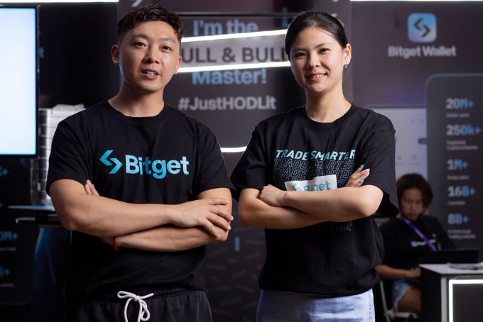 Two people wearing black Bitget branded t-shirts posing with crossed arms at a promotional event, with signage reading 'I'm the BULL & BULL Master!' and '#JustHODLit' visible in the background.