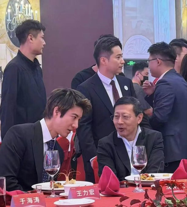 Several men in suits gather around a banquet table set with wine glasses and plates, with name placards in front of them reading '于海涛' and '王力宏'.