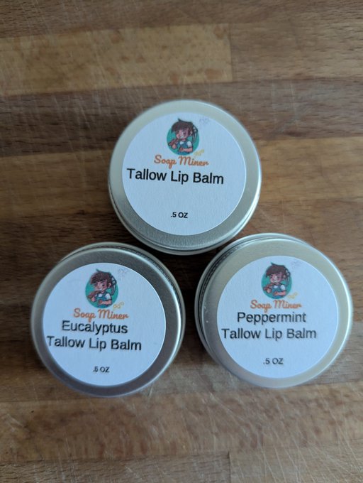 Three small 0.5 oz tins of 'Soap Miner' brand lip balm arranged on a wooden surface, featuring Tallow Lip Balm, Eucalyptus Tallow Lip Balm, and Peppermint Tallow Lip Balm varieties.