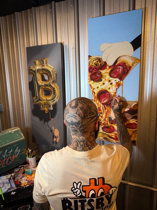 Photo from behind of a heavily tattooed person wearing a white "Bitsby" t-shirt, painting one of two canvases depicting a crypto-themed "B" balloon and a slice of pepperoni pizza being pulled away, in what appears to be an art studio with metal walls.