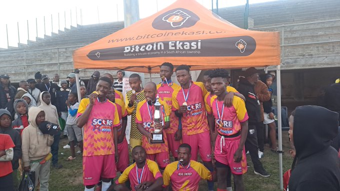 A sports team wearing pink and yellow 'SCORE' jerseys poses with a trophy under an orange 'Bitcoin Ekasi' tent at a stadium.