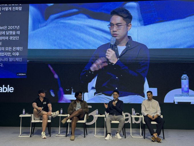 A panel discussion takes place at Korea Blockchain Week (KBW), with four speakers seated on stage while one is featured on a large screen behind them holding a microphone, consistent with the post's mention of speaking at the main stage thanks to 0G's invitation.