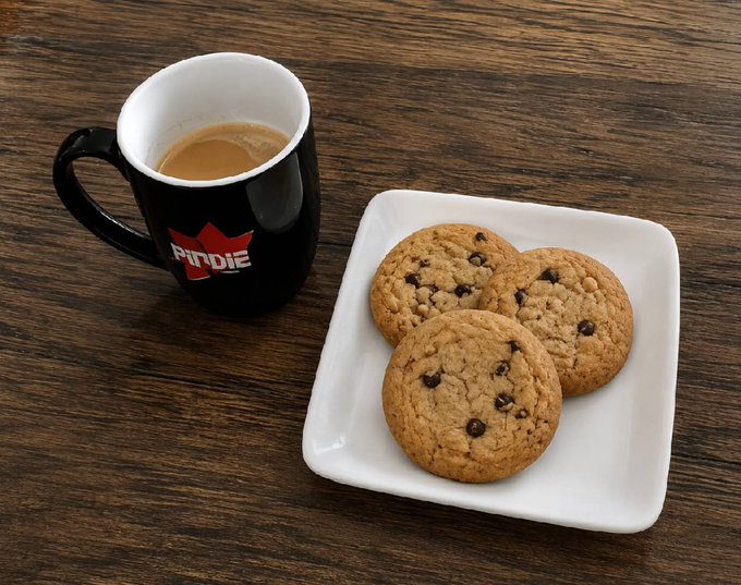 A black mug labeled "PIDDIE" filled with coffee sits next to a white square plate holding three chocolate chip cookies on a wooden table.