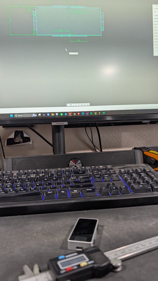 A desk setup featuring a computer monitor displaying a CAD drawing in green, a black mechanical keyboard with blue backlighting, and digital calipers resting on the desk.