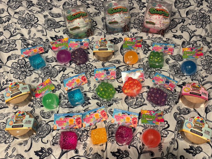 An assortment of colorful squishy toys in mesh packaging arranged on a black and white patterned bedspread.