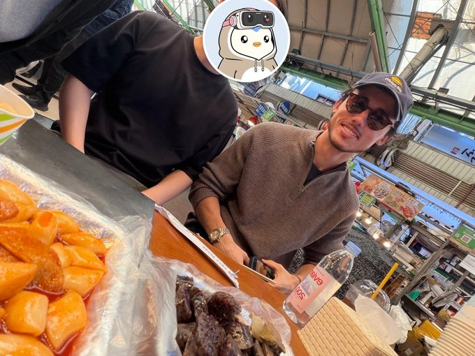 A casual market scene with two people at a food stall selling orange and dark-colored snacks, with one person's face covered by a cartoon animal sticker.